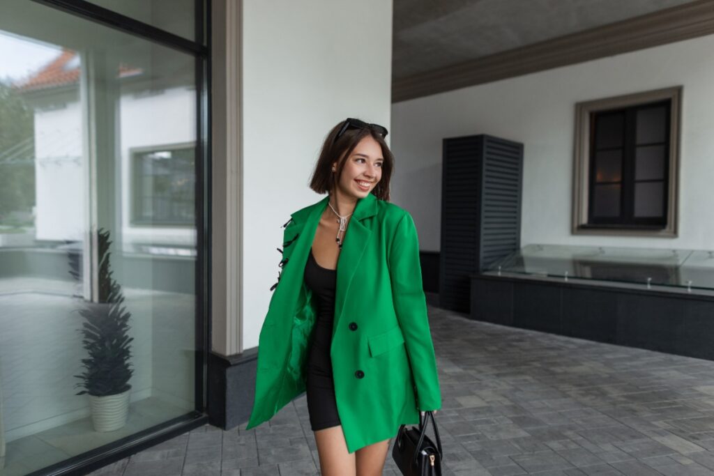 Une femme souriante avec une robe noire, un sac noir et un blazer vert